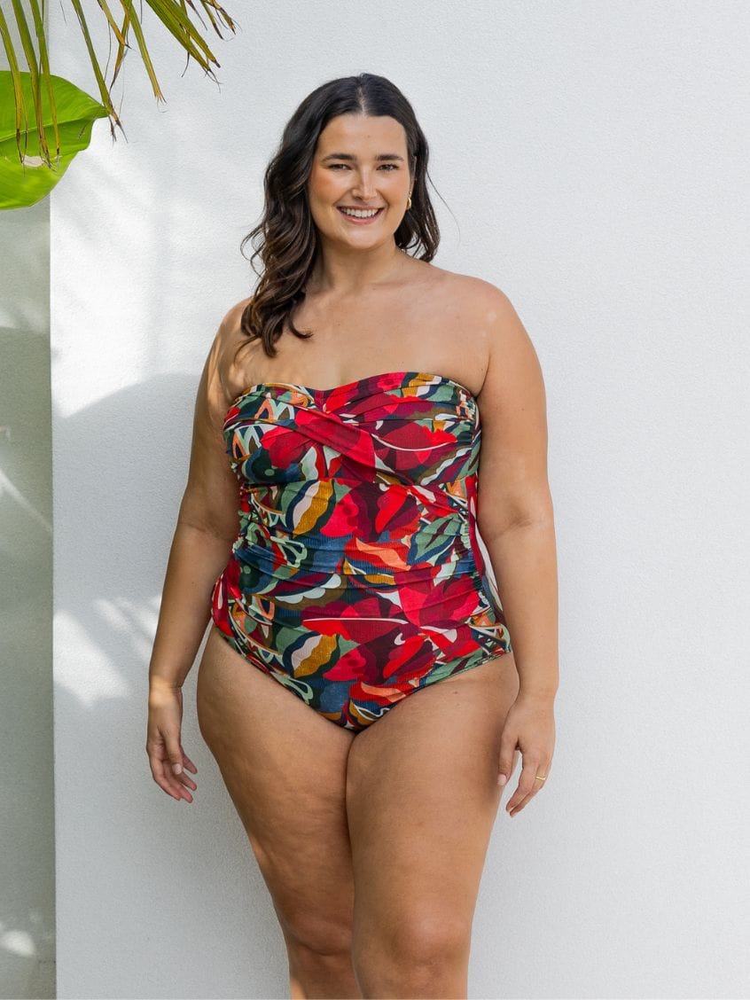 A woman modeling a strapless one-piece swimsuit with a vibrant tropical floral print in red, green, blue, and orange, standing against a white wall.
