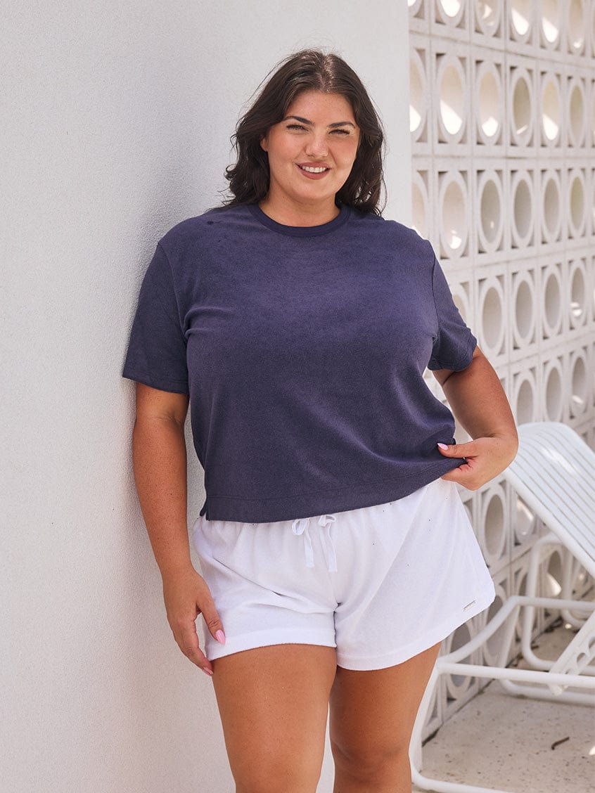 Woman wearing a navy blue t-shirt and white shorts standing against a white wall.