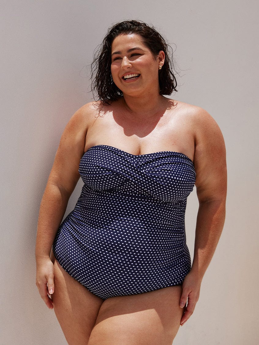 brunette model wearing a strapless one piece bathing suit with twist detail in between the breast area