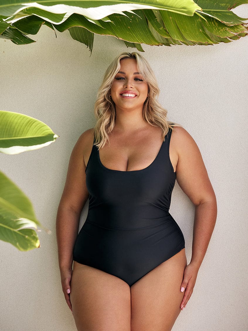 Women wearing a black one piece swimsuit standing in front of a white wall