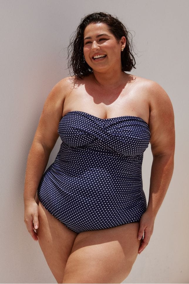 brunette model wearing a strapless one piece bathing suit with twist detail in between the breast area