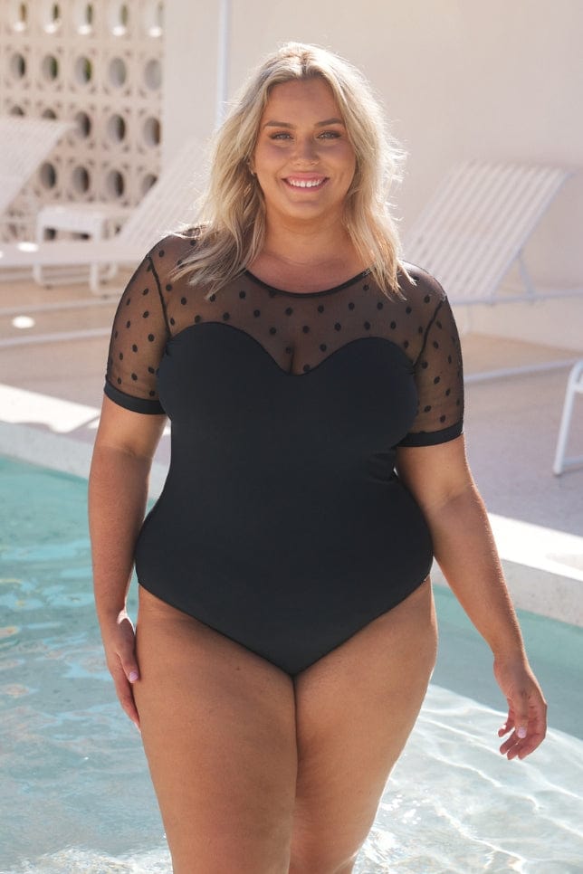 blonde model standing in front of a pool wearing a black sweetheart neckline one piece with mesh polkadot short sleeves