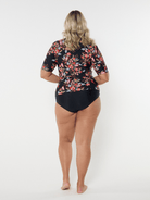 Woman wearing a floral rash guard and black swimsuit bottoms on a white background