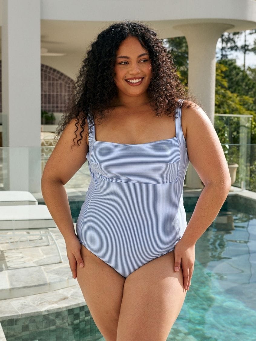 A woman modeling a light blue and white vertically striped one-piece swimsuit in front of a swimming pool and modern house patio.