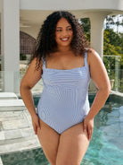 A woman modeling a light blue and white vertically striped one-piece swimsuit in front of a swimming pool and modern house patio.