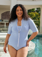 A woman with curly dark hair wearing a light blue and white vertically striped one-piece swimsuit with a front zipper, standing by a swimming pool.