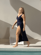 Woman in a navy swimsuit standing by a pool with a white lounge chair in the background.
