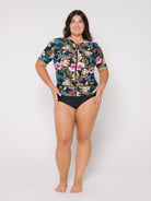 Woman wearing a floral rash guard and black swimsuit bottoms on a white background
