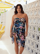 A woman models a strapless floral tube dress with a sweetheart neckline and a high side slit, set against a modern white block wall with circular cutouts