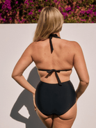A woman viewed from behind wearing a black one-piece high-waisted bikini with tie-back straps, standing against a white wall with purple flowers in the background.