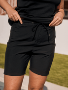 A person wearing black drawstring shorts and a black sleeveless top, standing outdoors with grass and a light wall in the background.