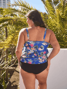 A woman shown from the back wearing a blue tropical floral print tankini swimsuit with black bottoms, standing outdoors in front of palm trees.
