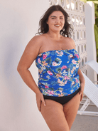 A woman modeling a strapless one-piece swimsuit with a vibrant blue floral top and solid black bottoms, standing against a white wall with a decorative screen.