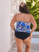 A woman viewed from behind wearing a strapless one-piece swimsuit with a blue floral top and solid black bottoms, standing outdoors next to a white decorative wall.