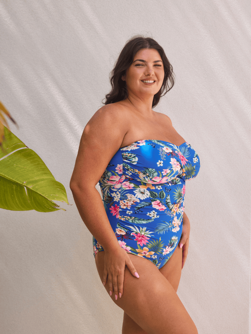 A woman modeling a strapless one-piece swimsuit with a bright blue background and a colorful tropical floral print, standing against a light beige textured backdrop with a green leaf on the left.