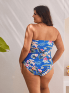 A woman wearing a strapless one-piece swimsuit with a vibrant blue tropical floral print, shown from the back against a white wall with soft shadows.