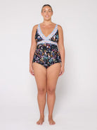 A woman modeling a one-piece swimsuit with a dark floral print and light blue crochet trim on a plain light gray studio background.