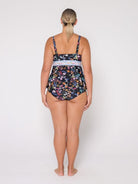 A woman stands with her back to the camera wearing a one-piece swimsuit with a dark navy floral print and a light blue patterned waistband, photographed against a plain white studio background.