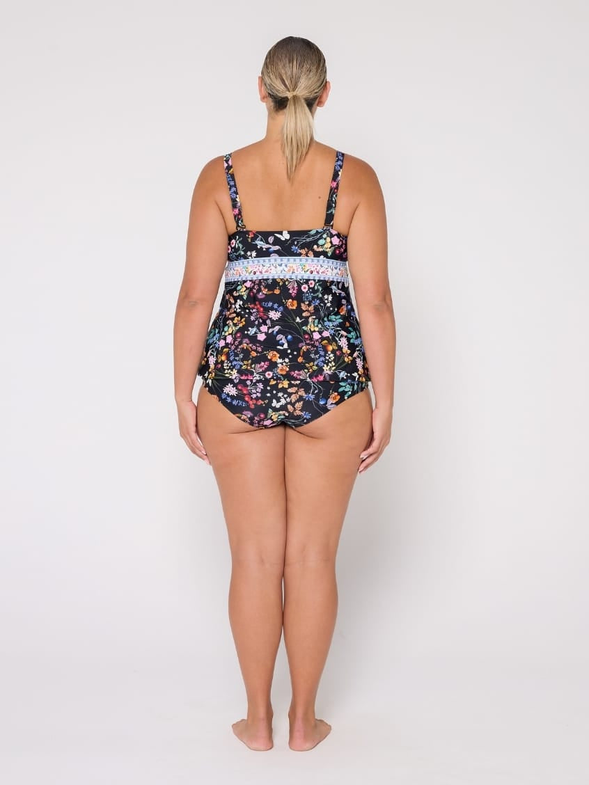 A woman stands with her back to the camera wearing a one-piece swimsuit with a dark navy floral print and a light blue patterned waistband, photographed against a plain white studio background.