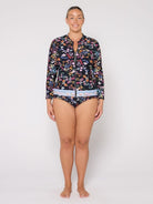 A woman modeling a two-piece swimsuit set consisting of a long-sleeve floral-print zip-front top and matching bikini bottoms, photographed against a plain light gray studio background.