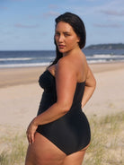 A woman wearing a black one-piece swimsuit stands on a sandy beach with the ocean and sky in the background, looking over her shoulder.