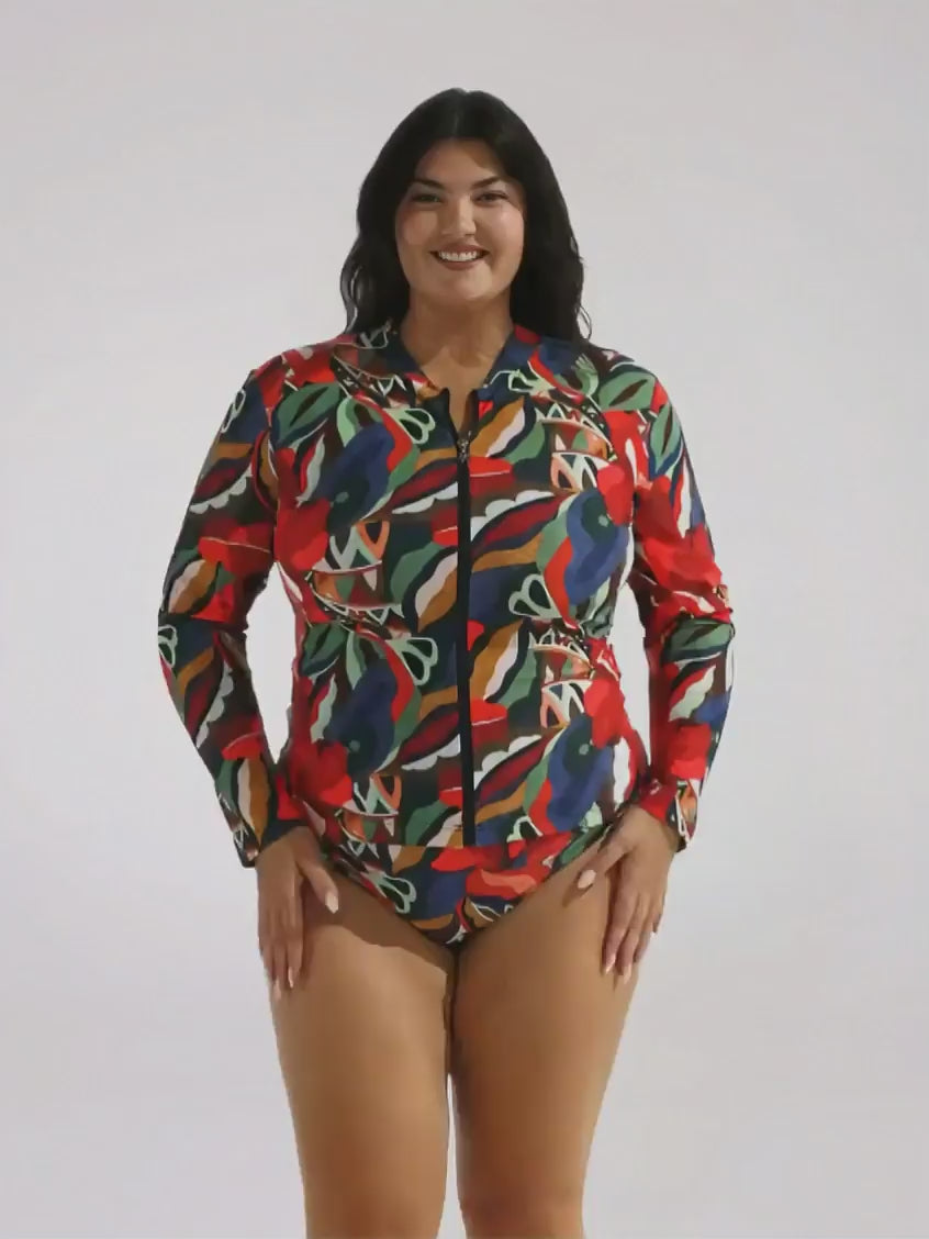 A woman modeling a long-sleeve, collared one-piece swimsuit with a colorful abstract print, standing against a plain light gray studio background.