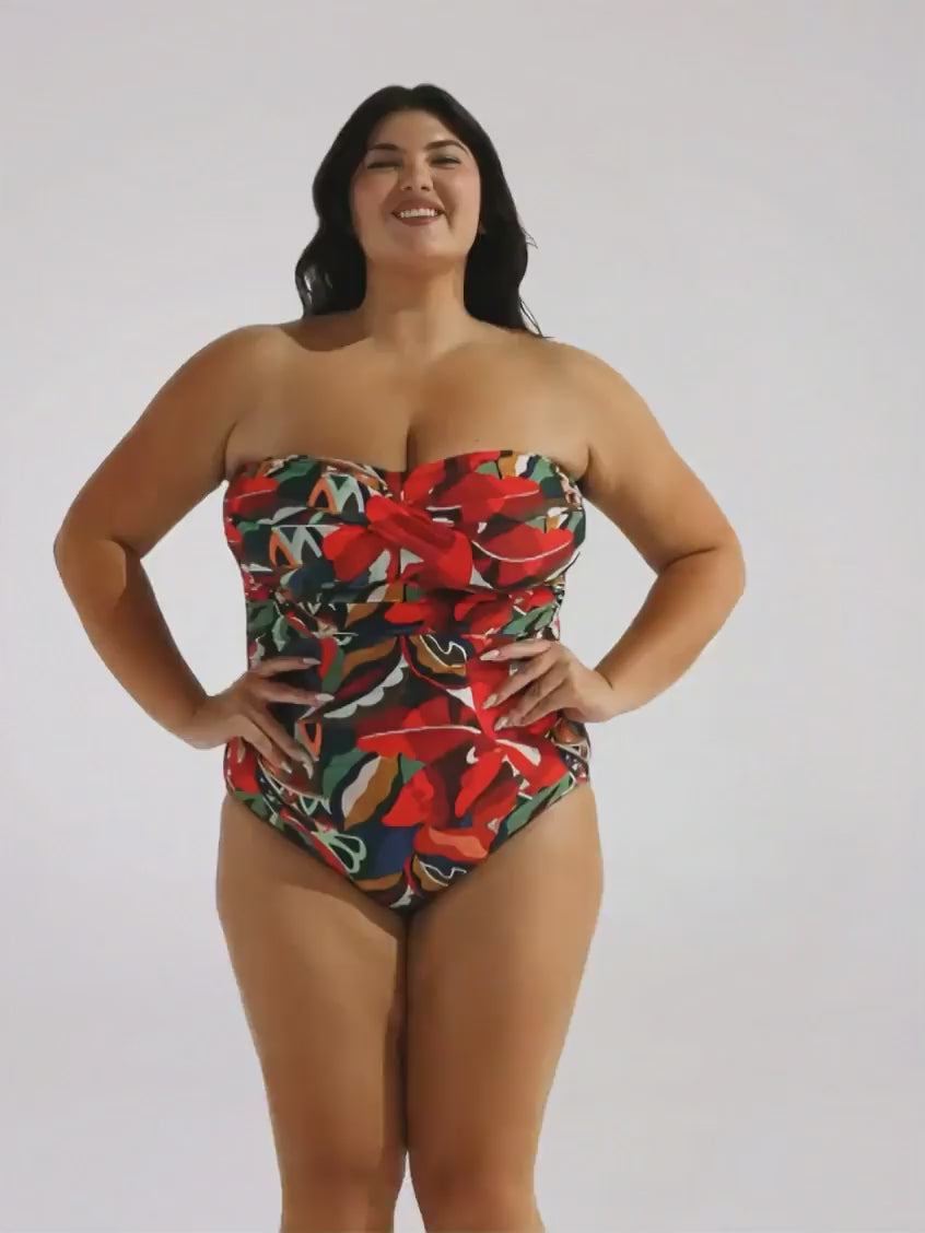 A woman modeling a strapless one-piece swimsuit with a vibrant tropical floral print in red, green, yellow, and blue, standing against a plain light gray studio background.