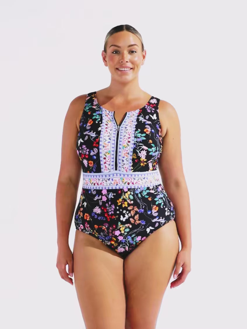 A woman modeling a one-piece swimsuit with a dark navy floral print and a light blue embroidered V-neck panel, standing by a swimming pool in front of a modern white building.