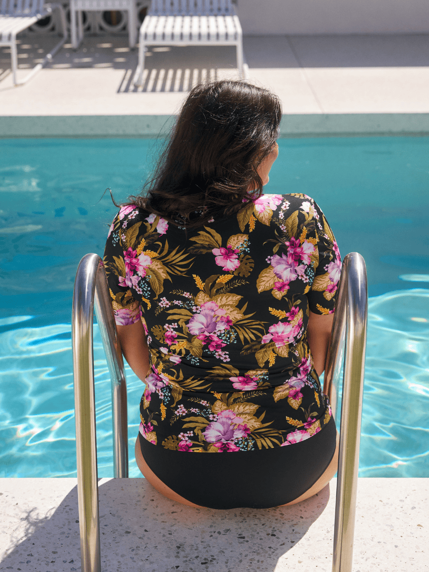 A woman viewed from behind sitting on the edge of a swimming pool, wearing a black short-sleeve floral top with pink, purple, and yellow tropical flowers, and black bottoms.