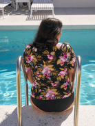 A woman viewed from behind sitting on the edge of a swimming pool, wearing a black short-sleeve floral top with pink, purple, and yellow tropical flowers, and black bottoms.
