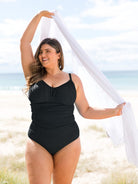 A woman stands on a sandy beach wearing a black one-piece ruched swimsuit and holding a white scarf overhead.