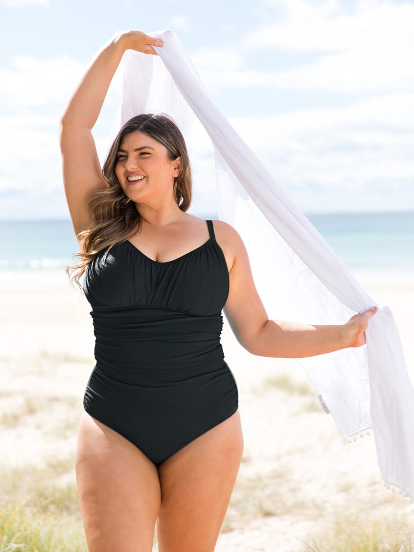 A woman stands on a sandy beach wearing a black one-piece ruched swimsuit and holding a white scarf overhead.